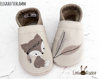 May include: A pair of light gray and taupe soft-soled baby shoes featuring an embroidered fox with a feather headband on the left shoe and two leaves on the right shoe. The interior is lined with taupe leather and includes a Little Rabbit brand label.