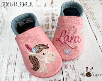 Op de afbeelding: Roze leren babyschoentjes met een paardenontwerp en de naam "Lara" geborduurd op één schoen. De schoentjes hebben een blauwe voering en een Little Rabbit logo.