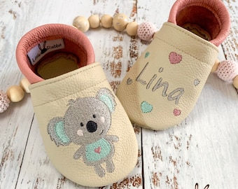 Op de afbeelding: Een paar beige leren babyschoentjes met roze bies. Eén schoen heeft een geborduurde koala en de andere schoen heeft de naam "Lina" geborduurd met roze en groene hartjes.