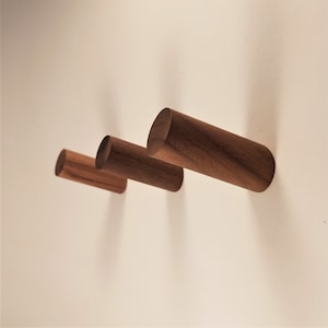 May include: Three wooden wall hooks in different sizes, made of dark brown wood. The hooks are mounted on a white wall.