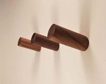 May include: Three wooden wall hooks in different sizes, made of dark brown wood. The hooks are mounted on a white wall.