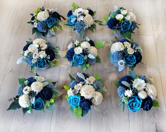 May include: Nine small floral arrangements in shades of blue, teal, and cream, with green foliage. The flowers are arranged in small, gray pots and sit on a light wood surface. The bouquets feature various flower types, including roses and other decorative blooms.