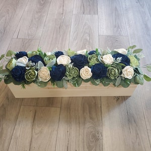 May include: A wooden box filled with a floral arrangement of blue, white, and green sola wood flowers. The flowers are arranged in a rectangular box with a natural wood finish.