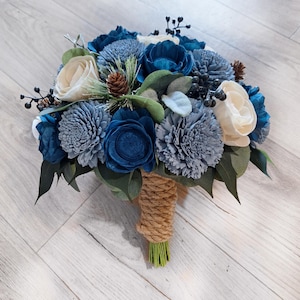 May include: A bouquet of blue, gray, and cream-colored flowers with green leaves and pinecones. The stems are wrapped in twine and green floral tape. The arrangement features various flower types and textures.