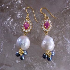 May include: A pair of gold-coloured drop earrings. Each earring features a cluster of small, dark blue pearls at the bottom, a large white pearl in the middle, and a ruby surrounded by clear crystals at the top. The earrings have a hook closure.