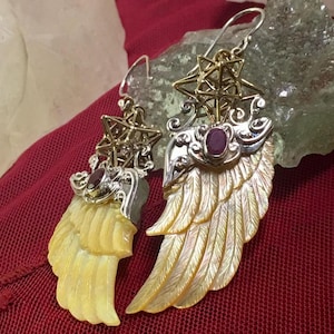 Puede incluir: Un par de pendientes de plata de ley con un tema celestial. Cada pendiente presenta un diseño de ala de nácar, acentuado con una gema de rubí y una estrella dorada. Los pendientes, con cierre de gancho, están sobre un fondo rojo.