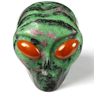 Op de afbeelding: Een groene alienhoofdsculptuur met rode en zwarte accenten. De alien heeft grote, ovale, oranjerode ogen. De sculptuur is gemaakt van een gespikkelde, gepolijste steen.
