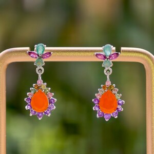 May include: A pair of drop earrings featuring a central pear-shaped orange stone surrounded by a halo of small purple and multi-colored gemstones. The top section includes a green stone flanked by two purple marquise-cut stones in silver settings.