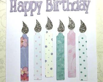 Może przedstawiać: Ręcznie robiona kartka urodzinowa z napisem "Happy Birthday" w lawendowych literach. Sześć kolorowych świec z błyszczącymi płomieniami jest ułożonych pod tekstem. Świece są ozdobione wzorami kwiatowymi i w kropki w odcieniach niebieskiego, różowego, zielonego i żółtego.