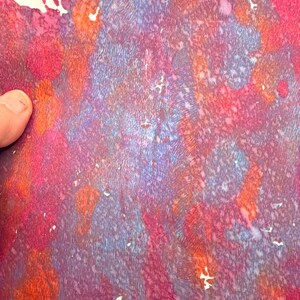 May include: Abstract artwork with a textured surface. The piece features a blend of colors, including shades of red, purple, orange, and blue. The artwork has a watercolor-like appearance with a mix of splatters and streaks.