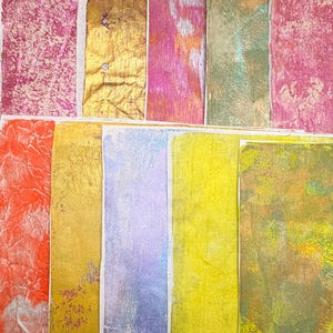 May include: An assortment of colorful, textured paper rectangles. The colors include shades of pink, gold, orange, green, yellow, and purple. The paper has a variety of textures, including crinkled and metallic finishes. The rectangles are arranged in a grid-like pattern.