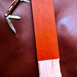 Puede incluir: Un cuchillo de bolsillo con mango marrón y blanco y tres hojas está junto a una piedra de afilar rectangular naranja. La piedra tiene una base de madera. Una correa de cuero está sujeta a la base de madera.