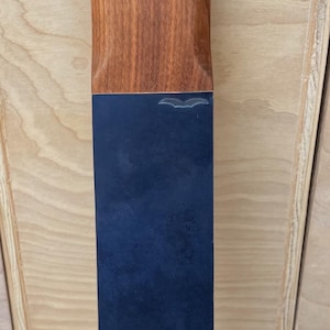 Puede incluir: Un tapón de botella rectangular con una parte superior de madera y un cuerpo azul oscuro. La parte superior de madera tiene una veta natural y un lazo de cuero para facilitar el manejo. El tapón está diseñado para adaptarse a una abertura de botella estándar.