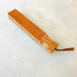 May include: A wooden razor strop with a leather surface. The strop features a light brown leather strip attached to a bamboo handle. A small leather loop with a brass clasp is attached to the handle.