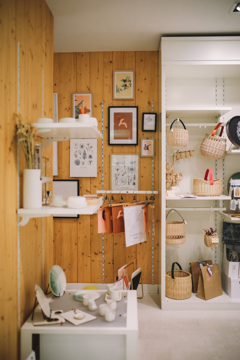 May include: A white shelf unit with various baskets and a brown paper bag. The shelves are filled with wicker baskets of different sizes and shapes. The baskets are hanging from the shelves and are empty. The shelves are white and the background is a light wood panel.