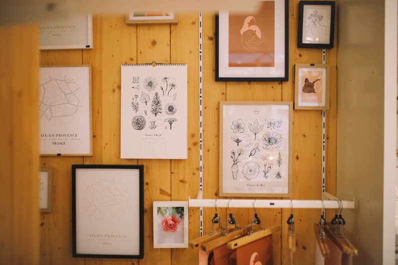 May include: A wall gallery featuring framed prints of botanical illustrations, maps, and abstract art. The prints are in black and white and feature a minimalist aesthetic. The wall is made of wood and has a light brown color.