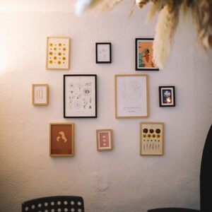 May include: A white wall with a collection of framed artwork and pressed flower displays. The artwork includes botanical illustrations, a map of Provence, France, and a portrait of a woman. The pressed flower displays feature various types of flowers in different colors.