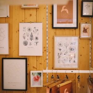 K&ouml;nnte beinhalten: Eine Wandgalerie mit gerahmten Drucken von botanischen Illustrationen, Karten und abstrakter Kunst. Die Drucke sind in Schwarzwei&szlig; gehalten und zeichnen sich durch eine minimalistische &Auml;sthetik aus. Die Wand ist aus Holz und hat eine hellbraune Farbe.