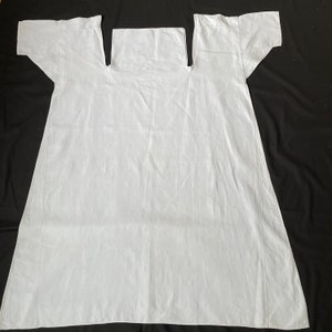 May include: A white linen garment with a square neckline and short sleeves. The garment is laid flat on a black surface.