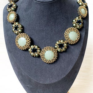 May include: A green and gold beaded necklace with round, light green stones. The necklace is displayed on a gray mannequin bust.