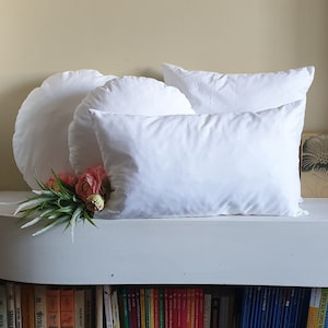 Puede incluir: Tres fundas de almohada blancas, dos redondas y una rectangular, dispuestas sobre una superficie blanca. La funda de almohada rectangular está encima de las dos fundas de almohada redondas. Un ramo de flores rosas se encuentra delante de las fundas de almohada.