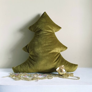 Puede incluir: Una almohada de árbol de Navidad de terciopelo verde con un adorno dorado. La almohada está sobre una superficie blanca con cuentas doradas.