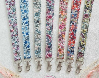 May include: A collection of seven floral patterned lanyards in various colors, including blue, pink, and orange. Each lanyard has a silver-colored metal clasp. The lanyards are arranged on a white surface.