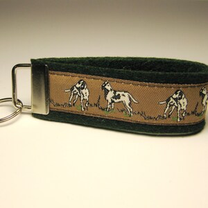May include: Green felt keyring with a brown ribbon featuring a repeating pattern of white and black dogs. The keyring has a silver metal ring.