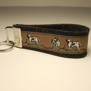 May include: Brown and tan key chain with a silver key ring. The key chain has a repeating pattern of black and white dogs.