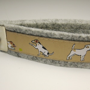 Può includere: Un portachiavi da polso in feltro grigio con un nastro beige con illustrazioni di cani. Il portachiavi ha un portachiavi in metallo color argento e una chiusura in metallo. Il nastro presenta immagini di cani in varie pose.