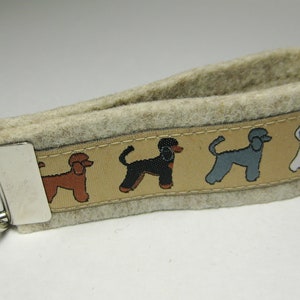 May include: A beige felt keyring with a silver key ring and a brown, tan, black, and white ribbon with a repeating pattern of poodle silhouettes.