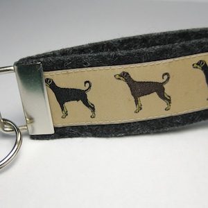May include: A grey felt keyring with a beige ribbon featuring three black and brown dog silhouettes. The keyring has a silver ring and a silver clasp.