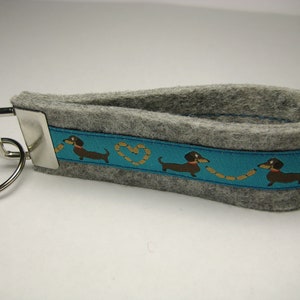 Op de afbeelding: Een grijze sleutelhanger van vilt met een blauw lint met een herhalend patroon van bruine teckels en gouden harten. De sleutelhanger heeft een zilveren sleutelring.