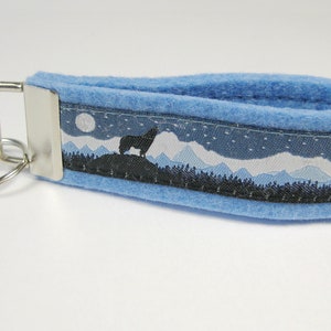 Könnte beinhalten: Ein blauer Schlüsselanhänger aus Filz mit einem silbernen Schlüsselring und einem schwarz-weißen Band mit einem Wolfssilhouetten, Bergen und einem Mond.