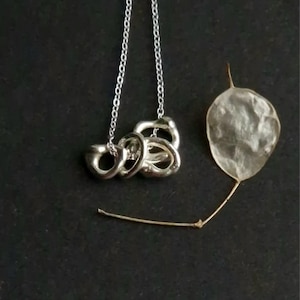 Peut inclure: Un collier argenté avec trois cercles entrelacés est présenté sur un fond sombre. Le pendentif est suspendu par une fine chaîne argentée. Une feuille sèche et translucide est placée à droite du collier, ajoutant une touche naturelle.