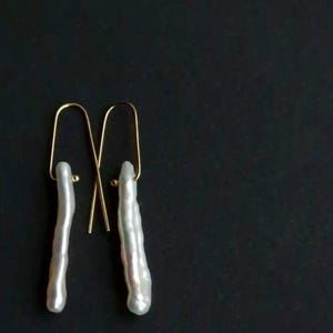 May include: A pair of drop earrings featuring long, slender, white iridescent stick pearls suspended from thin, gold-toned metal U-shaped wires. The pearls have a textured, organic surface with a subtle luster, hanging vertically from the simple gold hooks.