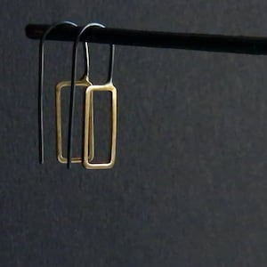 May include: A pair of gold-colored rectangular earrings hanging from a black rod against a dark background. The earrings have a simple, open frame design. The hooks are a dark metal.