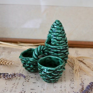 Peut inclure: Un ensemble de quatre contenants en céramique de couleur verte en forme de cône de pin. Les contenants sont empilés les uns sur les autres, le plus grand en bas et le plus petit en haut. Les contenants sont décorés d'un motif texturé de cône de pin.