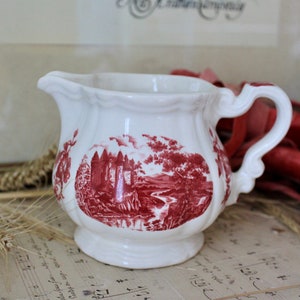 Puede incluir: Una jarra de crema de cerámica blanca con un patrón floral rojo y blanco. La jarra tiene un asa y un pico. Está sentada sobre un papel de música.