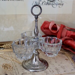 Peut inclure: Porte-condiments en argent avec trois coupelles en verre. Le porte-condiments a une poignée en forme d'anneau et une base ronde. Les coupelles sont en verre transparent avec un design facetté.