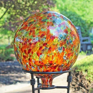 May include: A spherical glass gazing ball featuring a crackled texture with a vibrant mosaic pattern of red, orange, and yellow fragments. The orb is supported by a black metal stand with twisted wire accents at the base of the glass sphere.
