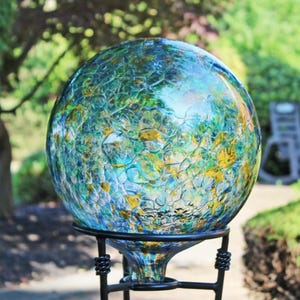 May include: A spherical glass gazing ball featuring a crackled texture with a blend of blue, green, and amber yellow colors. The orb is supported by a black metal stand with a circular base and twisted decorative accents on the vertical supports.