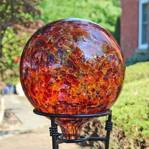 May include: A spherical glass gazing ball featuring a crackled texture with a vibrant mix of red, orange, and yellow hues. The orb is supported by a black metal stand with a circular ring base and decorative coiled wire accents at the top.