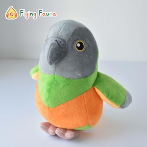 May include: A plush bird toy with a gray head, green chest, and orange belly. The toy has yellow eyes and a gray beak. The brand name "Flying Fauna" is visible in the upper left corner.