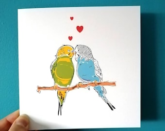 Pode incluir: Um cartão de felicitações com um desenho de dois periquitos, um verde e amarelo e o outro azul, empoleirados em um galho e se beijando. Há quatro corações vermelhos acima dos pássaros.