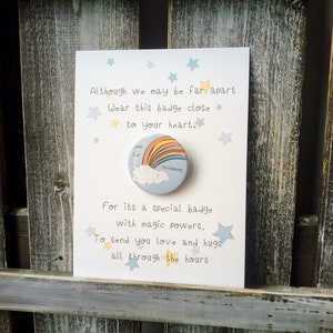 May include: A white card with blue stars and the text "Although we may be far apart Wear this badge close to your heart." A blue button with a rainbow and cloud design and the text "Look for Rainbows" is attached to the card. The text "For its a special badge with magic powers, To send you love and hugs all through the hours" is also on the card.