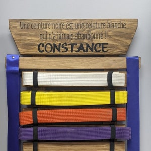 May include: A wooden belt rack with the text "Une ceinture noire est une ceinture blanche qui n'a jamais abandonné! CONSTANCE" on the top. The rack has five belts hanging on it, including a white belt, a yellow belt, an orange belt, a purple belt, and a blue belt. There are two medals hanging on the sides of the rack.