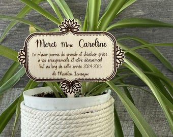May include: A wooden sign with a brown background and white text that reads "Merci Mme Caroline De m'avoir permis de grandir et d'évoluer grâce à vos enseignements et votre dévouement tout au long de cette année 2024-2025 de Marilou Levesque". The sign is sitting in a white pot with a brown and white rope wrapped around it.