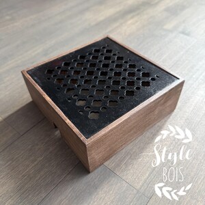 Peut inclure: Une boîte carrée en bois avec un motif noir découpé au laser sur le dessus. Le motif présente un motif répétitif de petits carrés et de cercles. La boîte est posée sur une surface en bois.