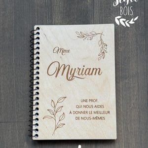 May include: A light wooden spiral-bound notebook with the name "Myriam" engraved on the cover. The cover also features engraved floral designs and the text "UNE PROF, QUI NOUS AIDES À DONNER LE MEILLEUR DE NOUS-MÊMES" and the number "#1".